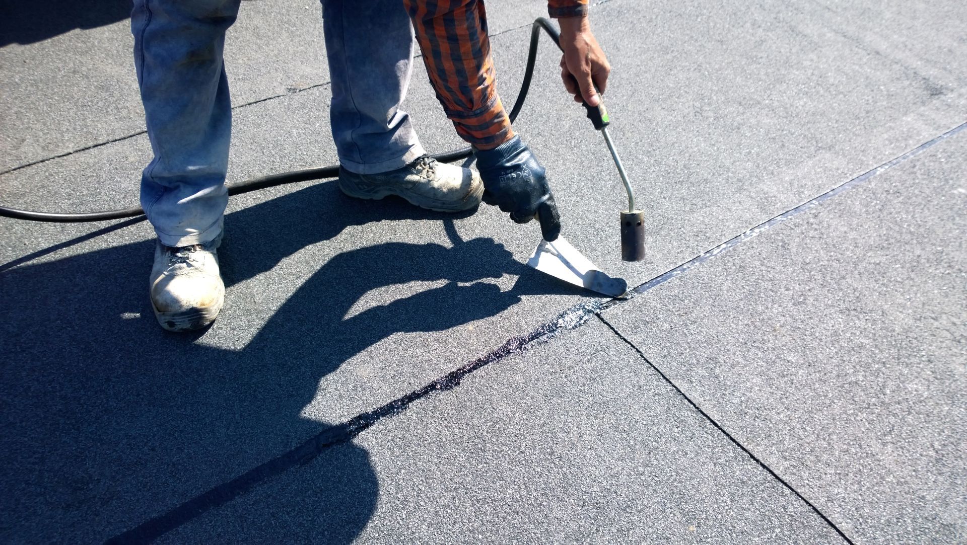 Worker sealing a roof seam with a torch on dark gray shingles
