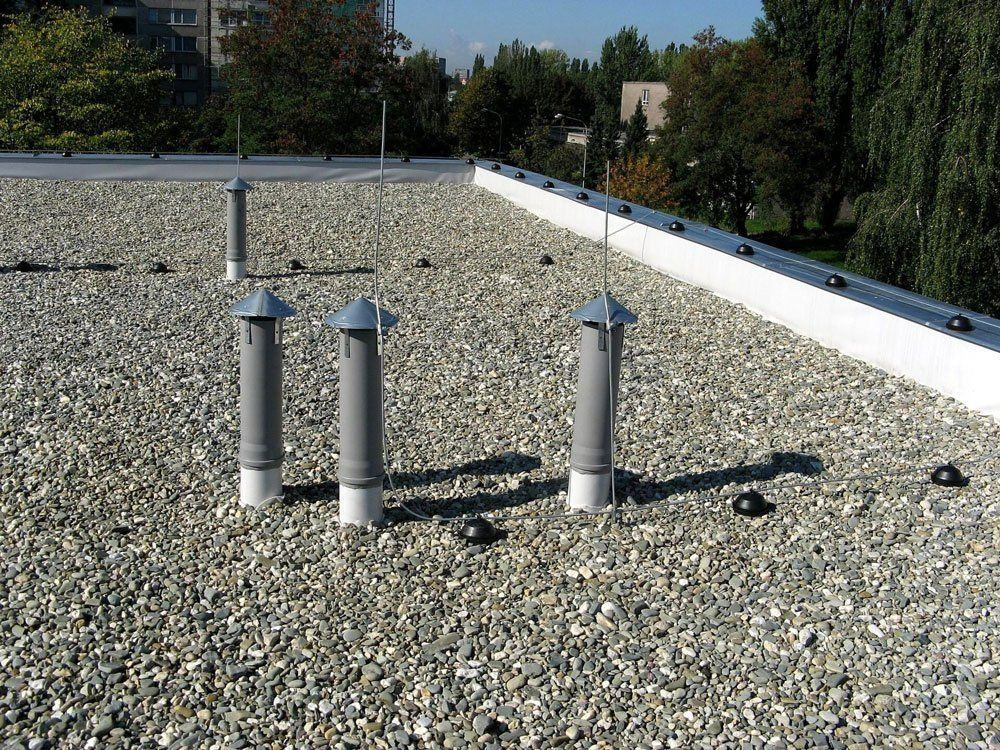 Gravel rooftop with three silver vent pipes and a white parapet under a clear sky