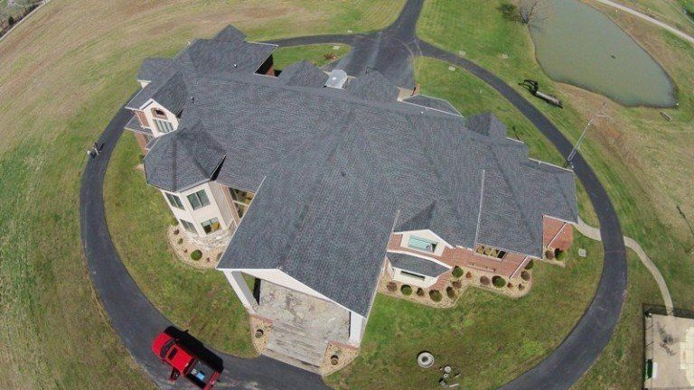Aerial view of a large house with a dark roof, curved driveway, and a red car parked nearby.