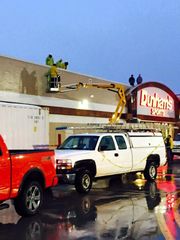 Workers on a ladder repairing a building roof above parked trucks at a Dillard's store in the rain