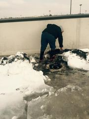 Person bending over a burned object in snow beside a concrete wall.