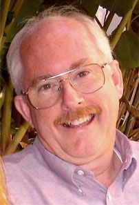 A picture of Michael Quinn. He is middle aged, with a big smile, white hair, brown moustache and square rim glasses.