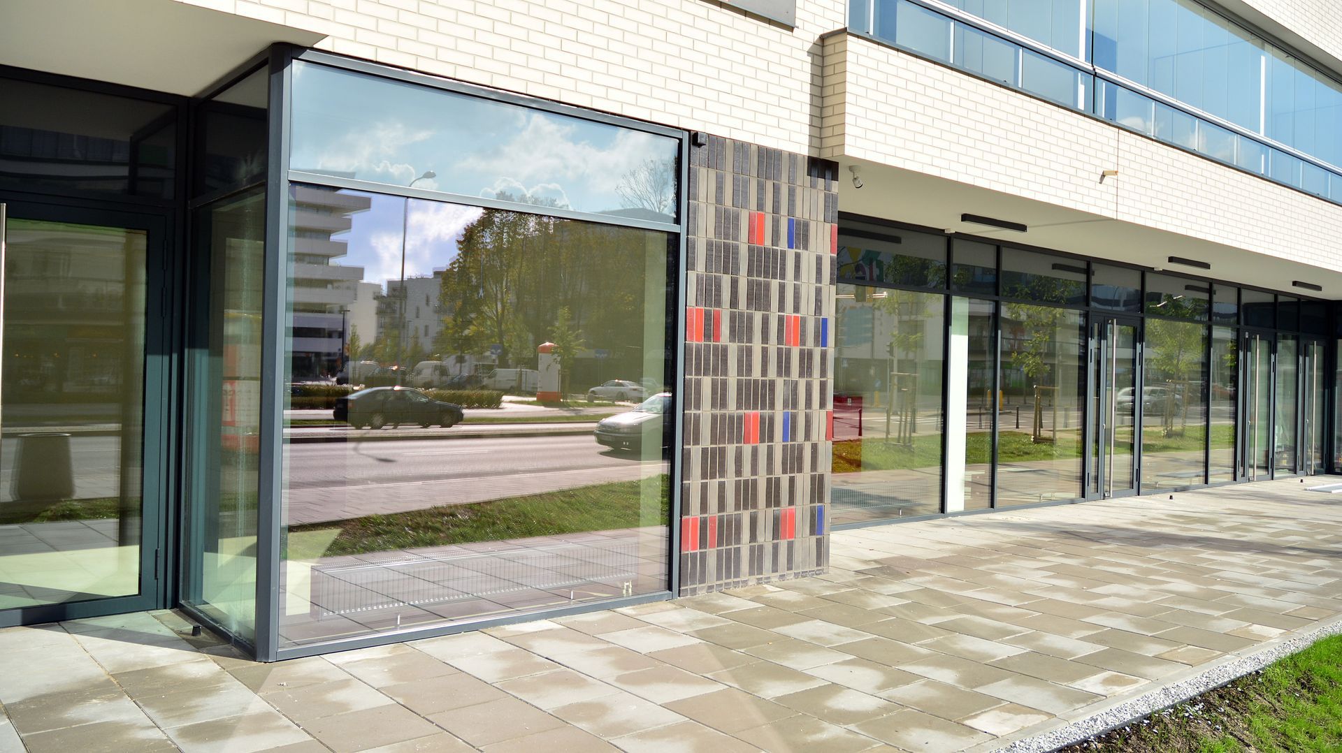 Modern storefront with large glass windows, dark frames, and a patterned brick column along a paved walkway.