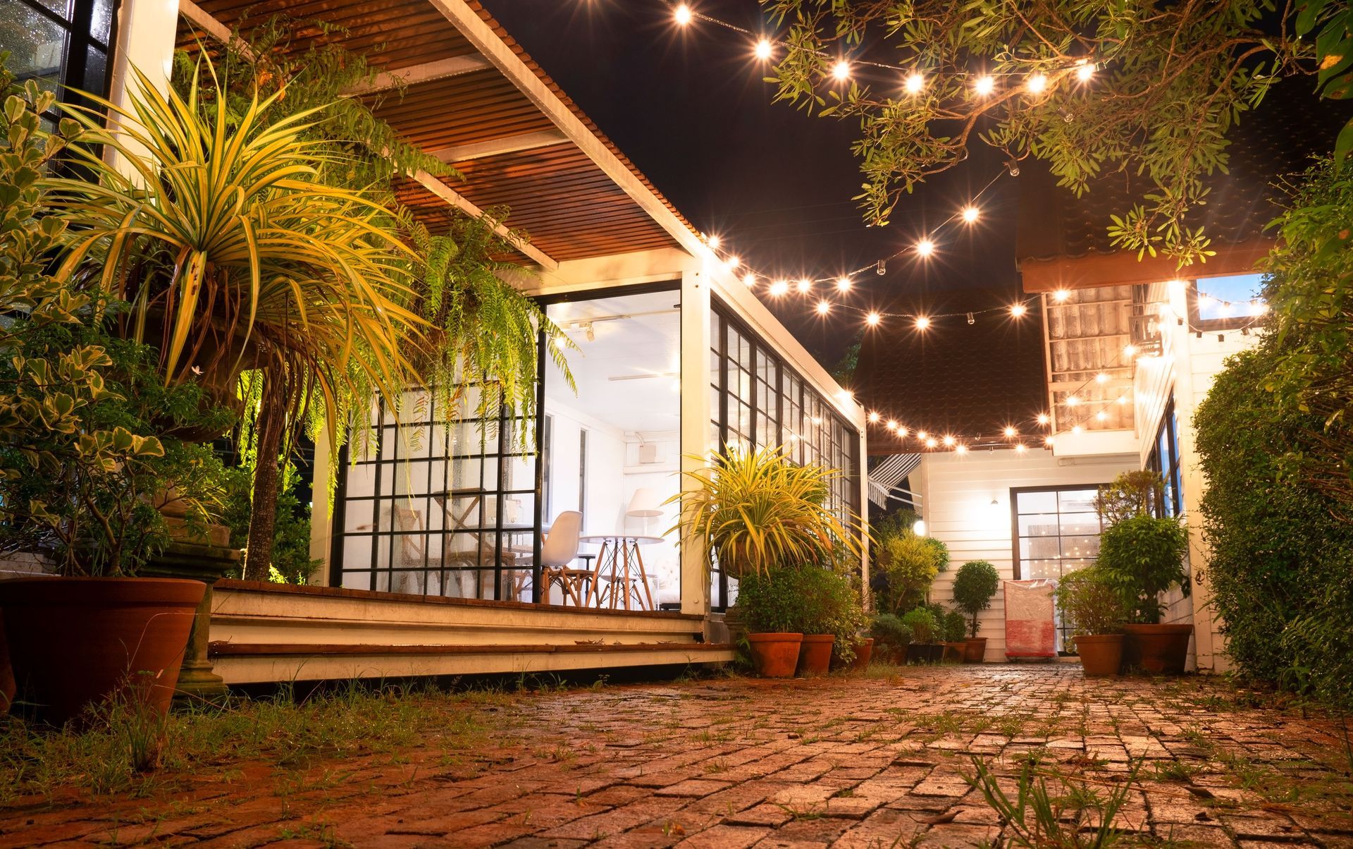 String lights illuminate a cozy garden patio with potted plants, a brick path, and a lit building at night