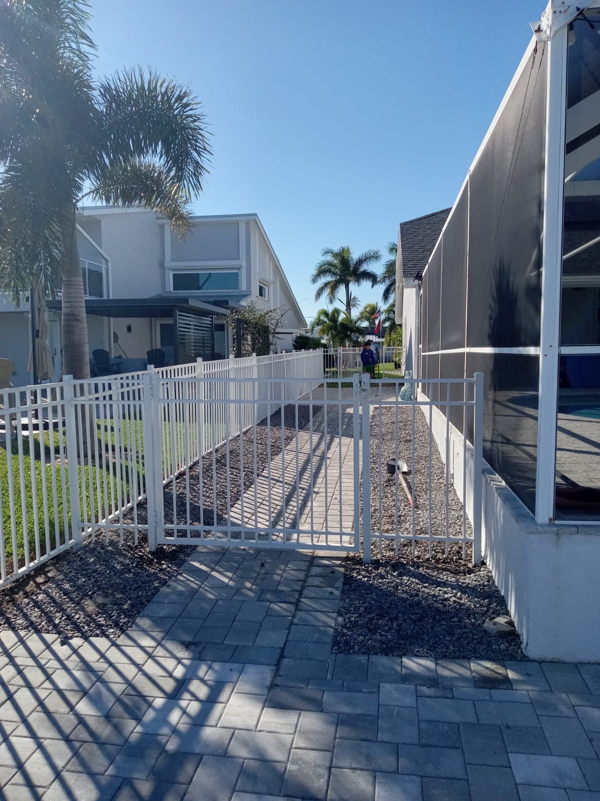 White fence and gate along a stone path, leading between houses under a blue sky.