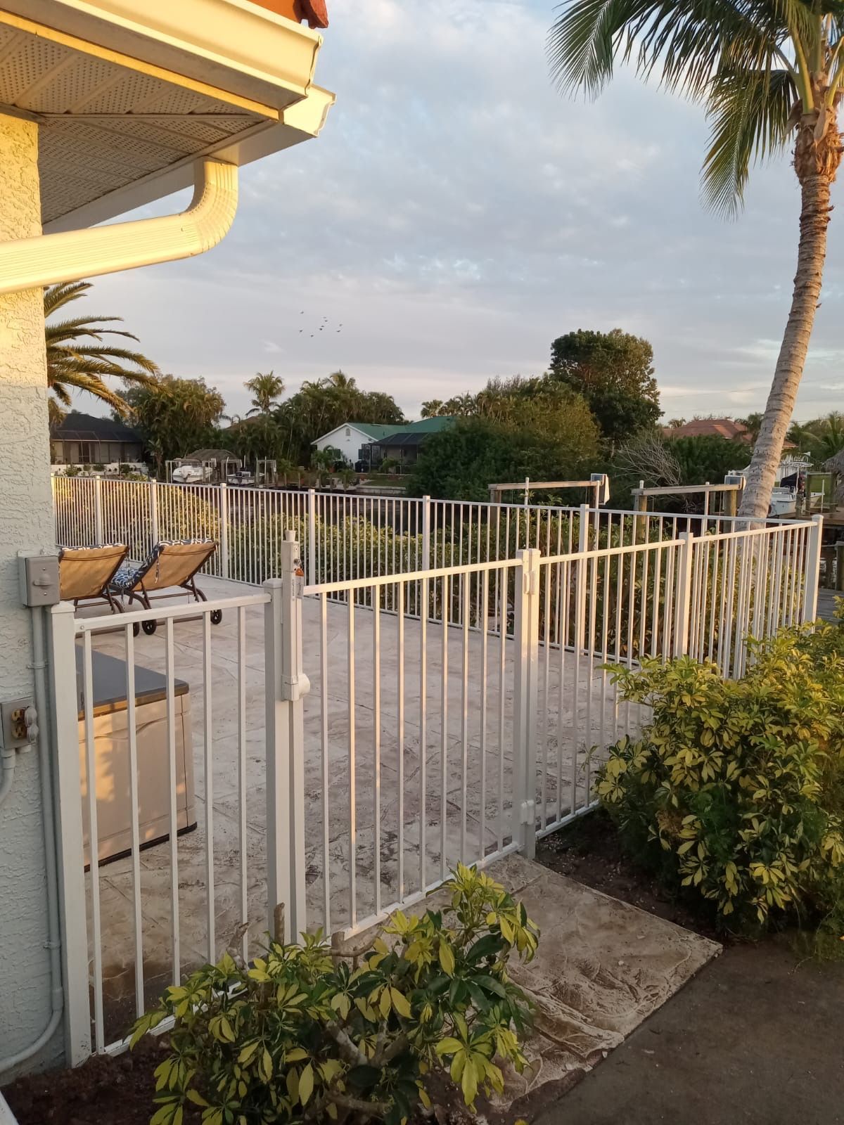 White fenced-in patio area with lounge chairs, next to a building and greenery, under a cloudy sky.