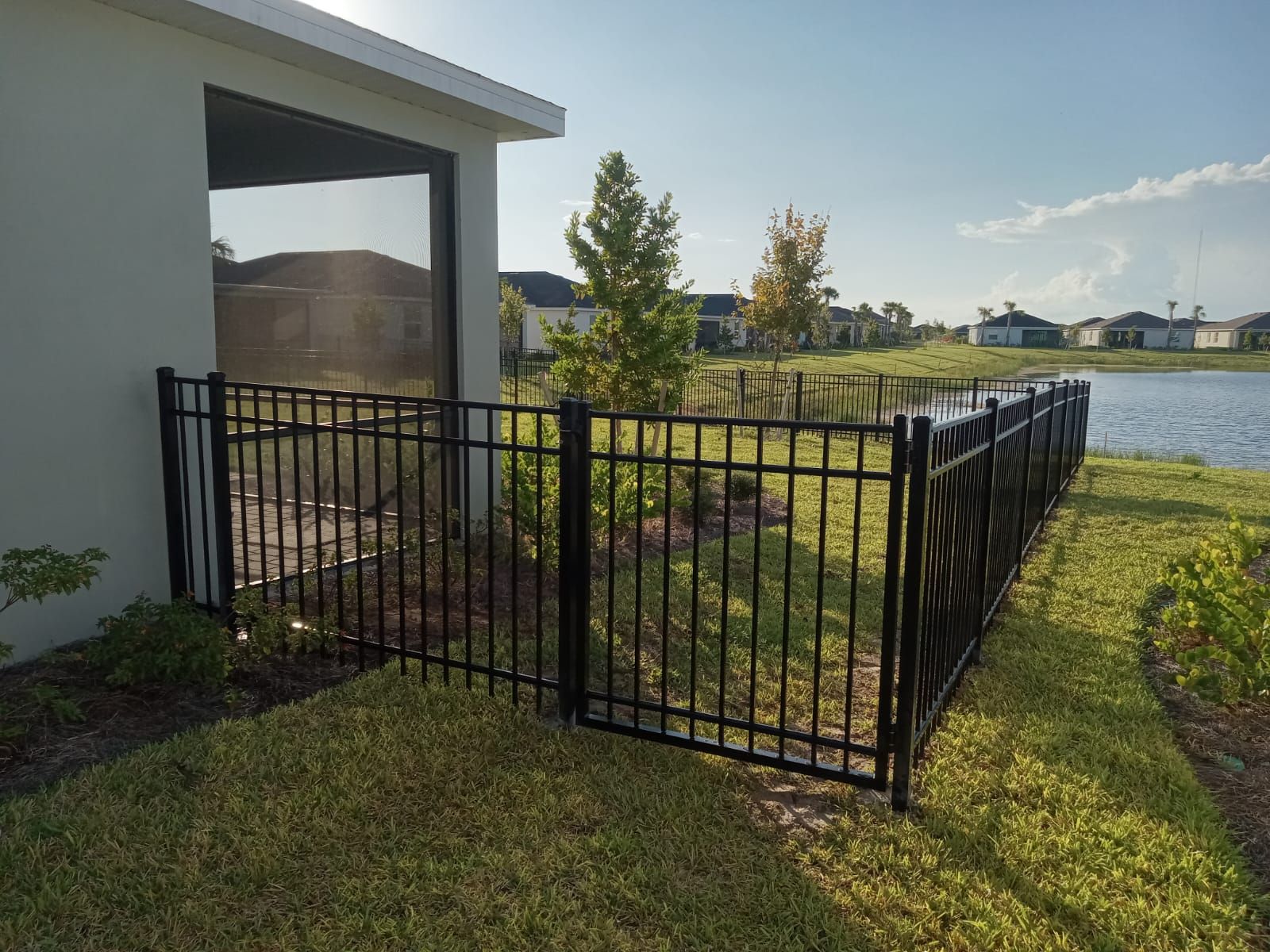 Black metal fence bordering a grassy yard next to a lake, with a house in the background.