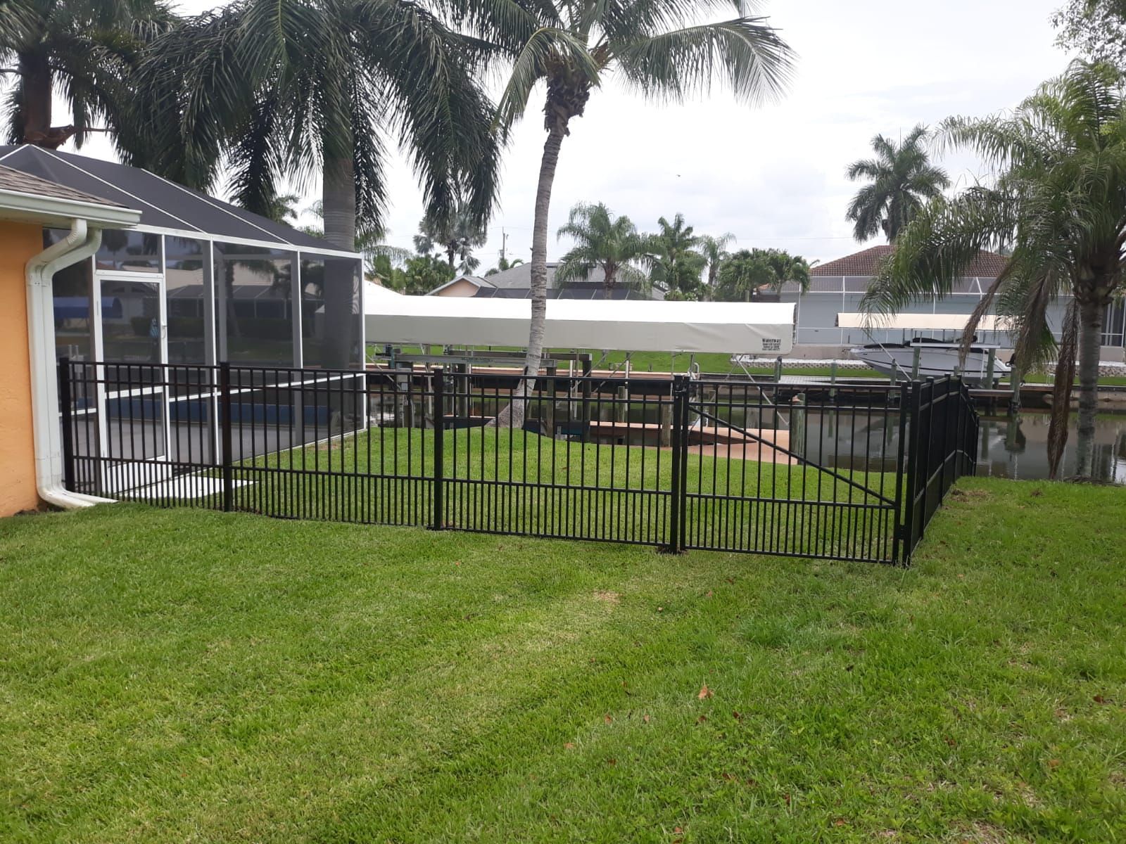 Black metal fence surrounds a screened patio and yard with canal in the background. Green grass and palm trees.