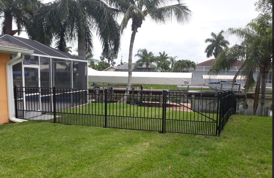Black fence surrounds a grassy yard leading to a canal with palm trees and a screened-in porch.