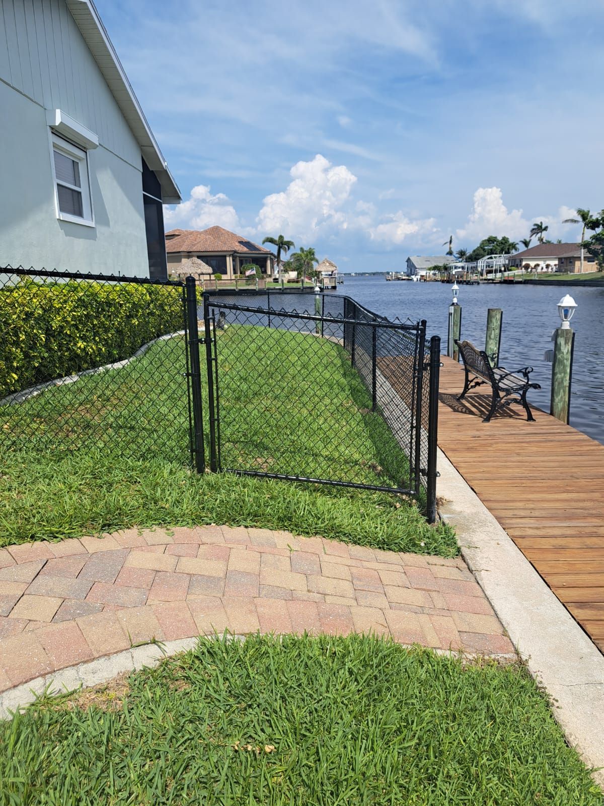 Black fence bordering a grassy yard beside a dock on a canal. Blue sky with clouds.