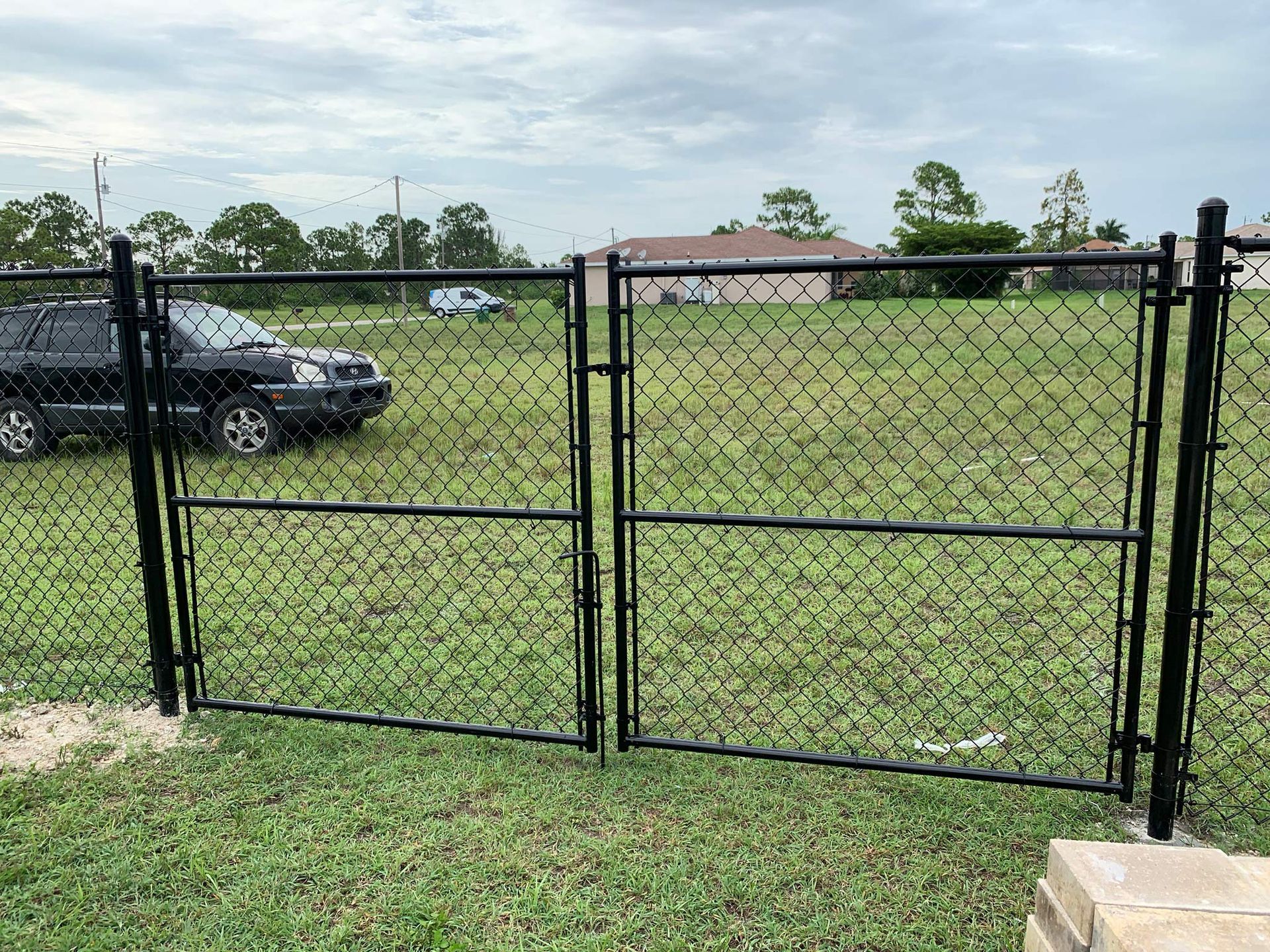 Black chain-link fence with double gates in a grassy yard, a car parked nearby. Cloudy sky.