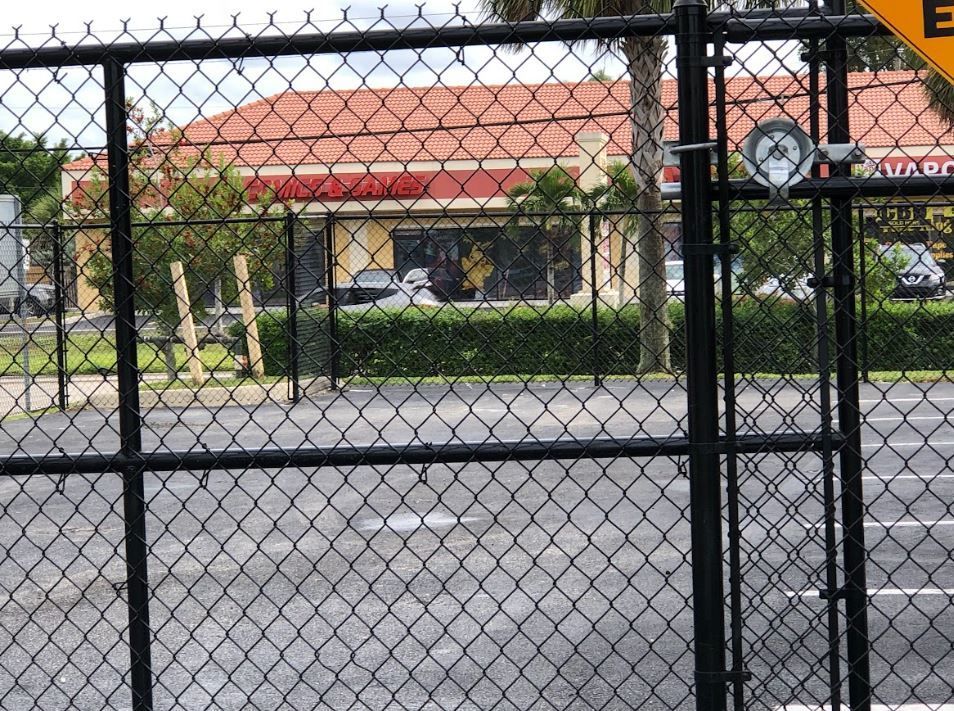 Black chain-link fence with a building in the background. The building has a red roof, and green bushes are visible.
