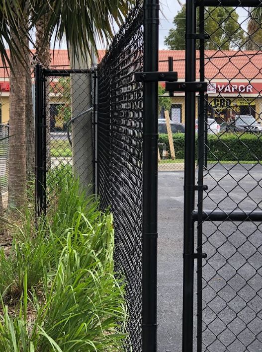 Black chain-link fence with a gate. Tall grass in foreground, buildings in background.