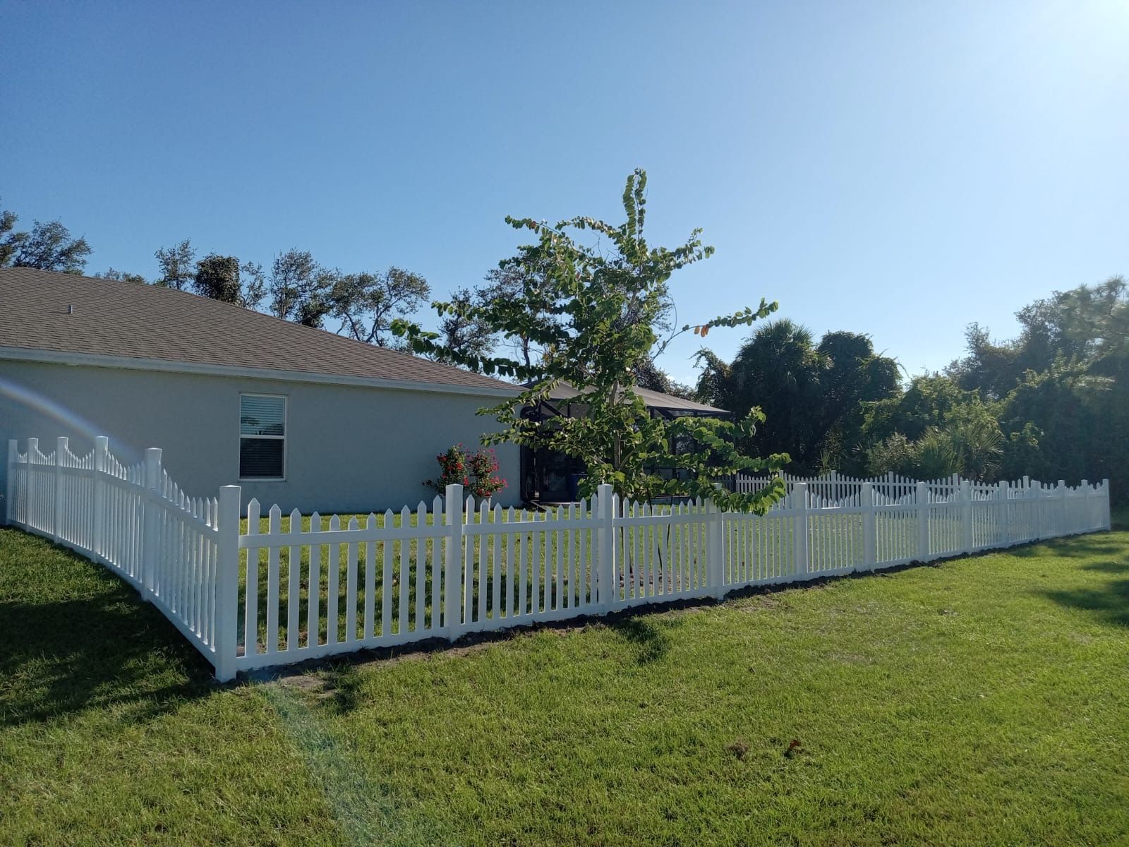 White picket fence borders green grass next to a house under a clear, blue sky.