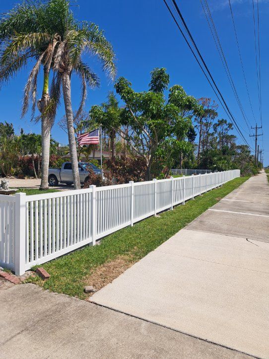 White picket fence lines a green lawn next to a sidewalk and road on a sunny day.