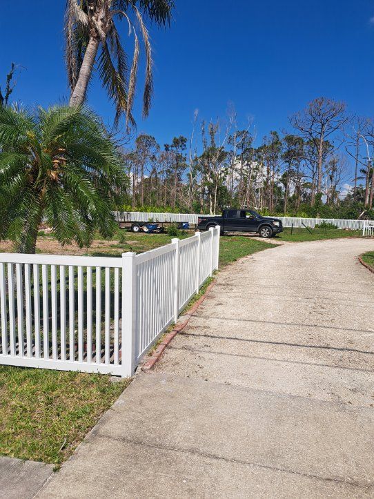 White fence, driveway, and black truck with a trailer on a sunny day. Trees in background.