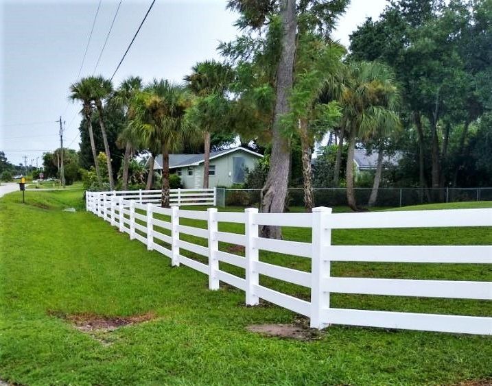 White picket fence borders a green lawn next to a road, with a blue house and palm trees in the background.