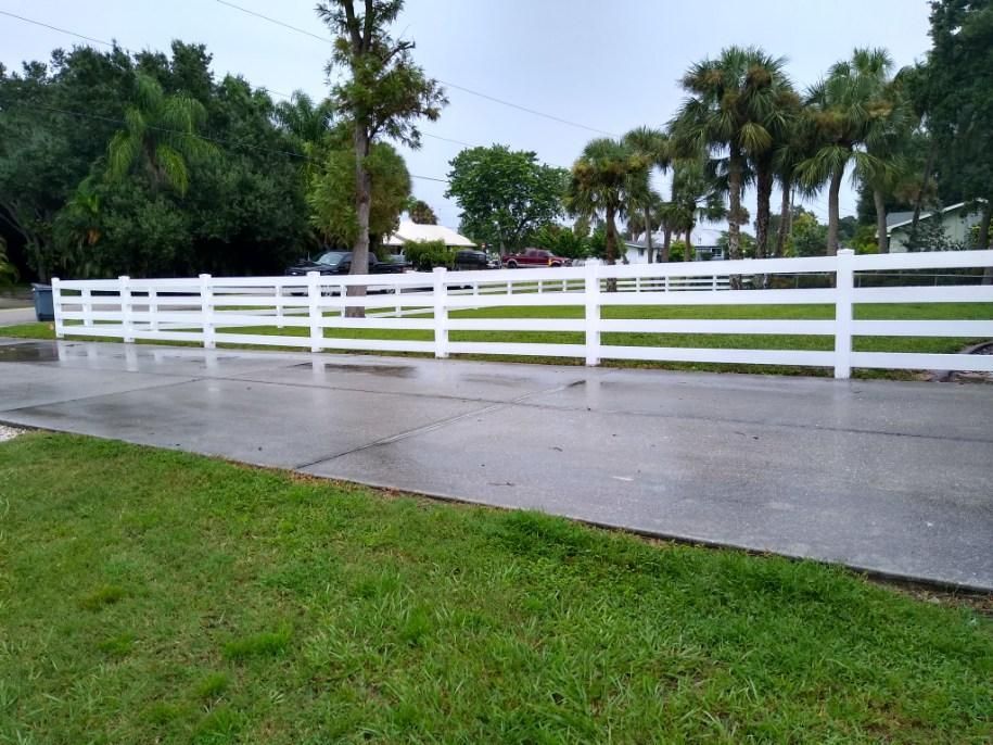 White fence along a wet road with green grass in the foreground and trees in the background.