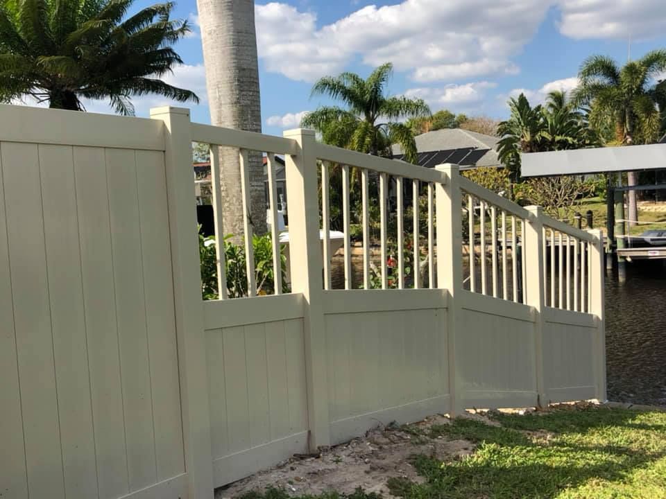 Beige vinyl fence near water with open top section. Palm trees in background under a blue sky.