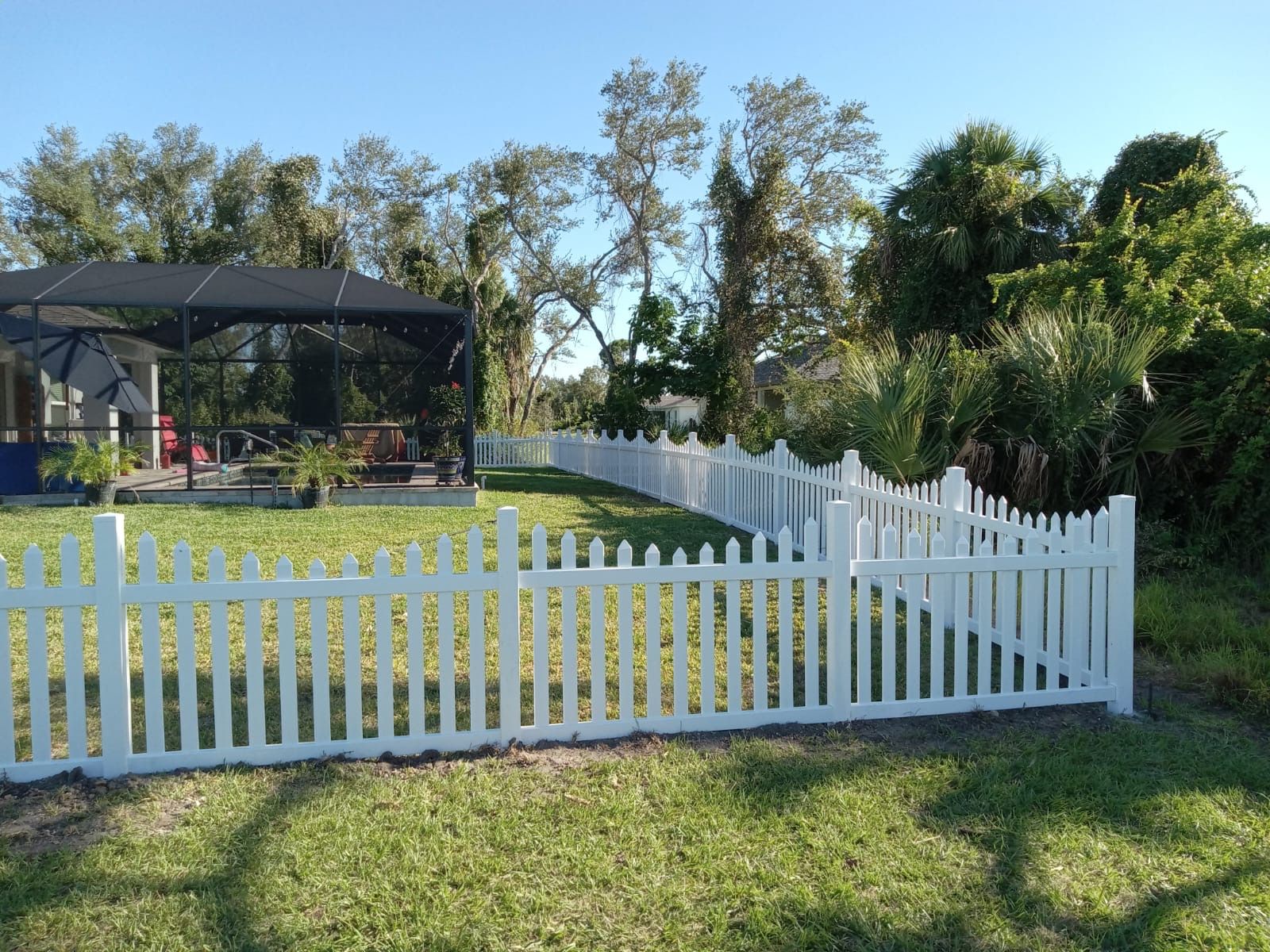 White picket fence borders a green yard, trees in the background, a house with a screen enclosure on the left.