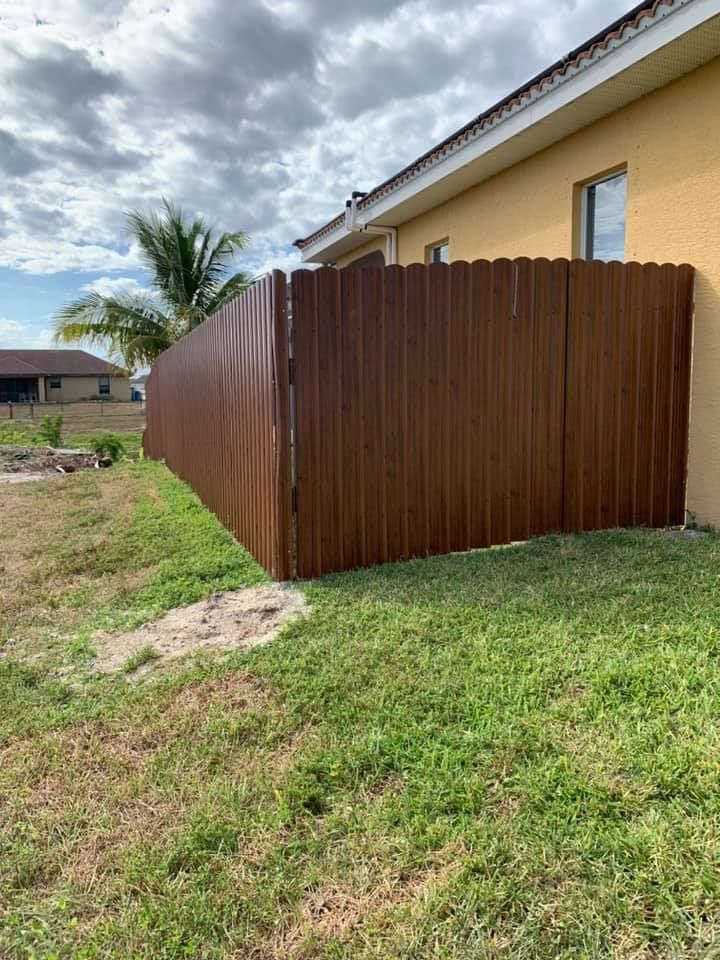 Brown fence next to a yellow house with a cloudy sky. Green grass in the foreground.