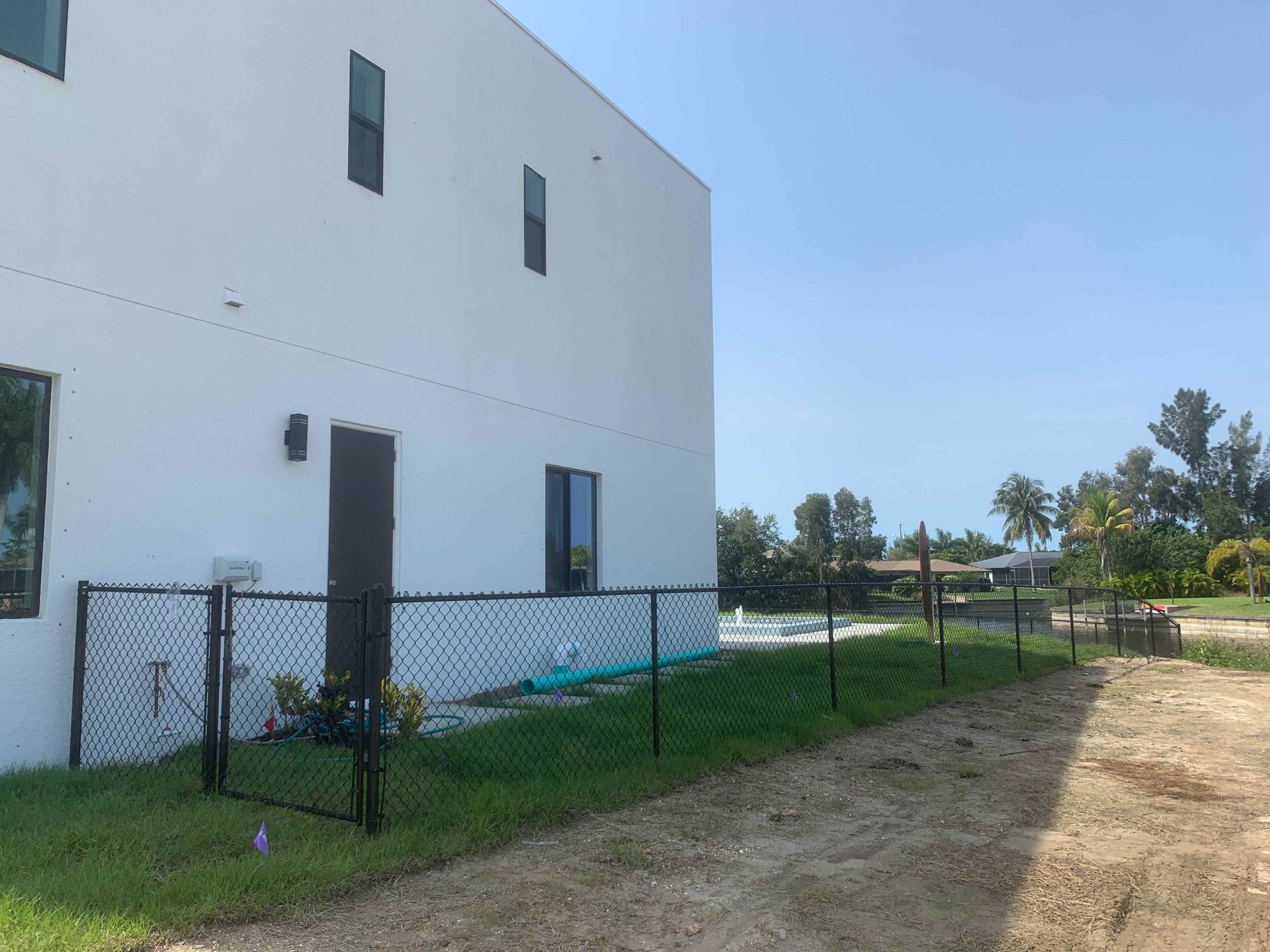 White building with black fence in front. Grassy area and dirt path. Blue sky.
