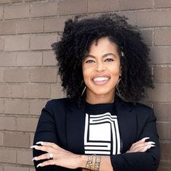 a woman with curly hair is standing in front of a brick wall with her arms crossed and smiling .