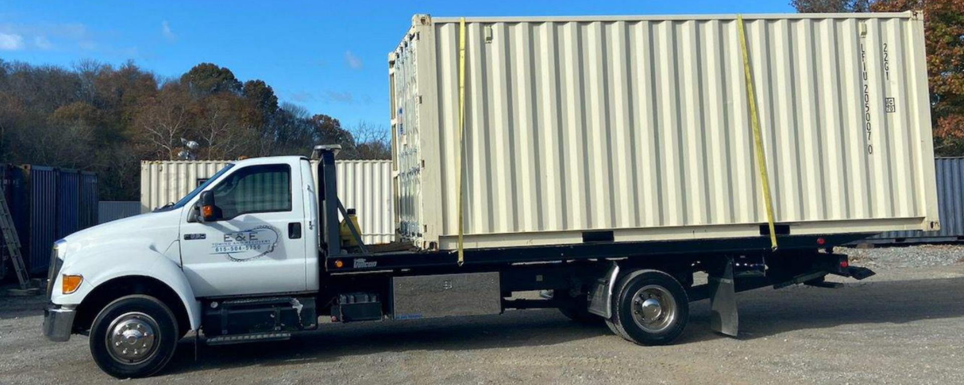 White truck carrying a large beige shipping container outdoors on a sunny day.