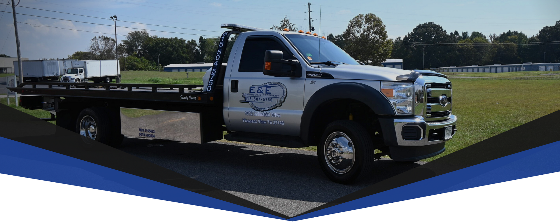 A silver tow truck with a flatbed is parked on asphalt with a field in the background.