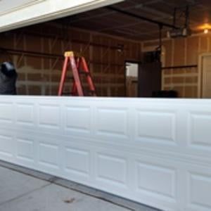 A white garage door is being installed in a garage with a ladder in the background.