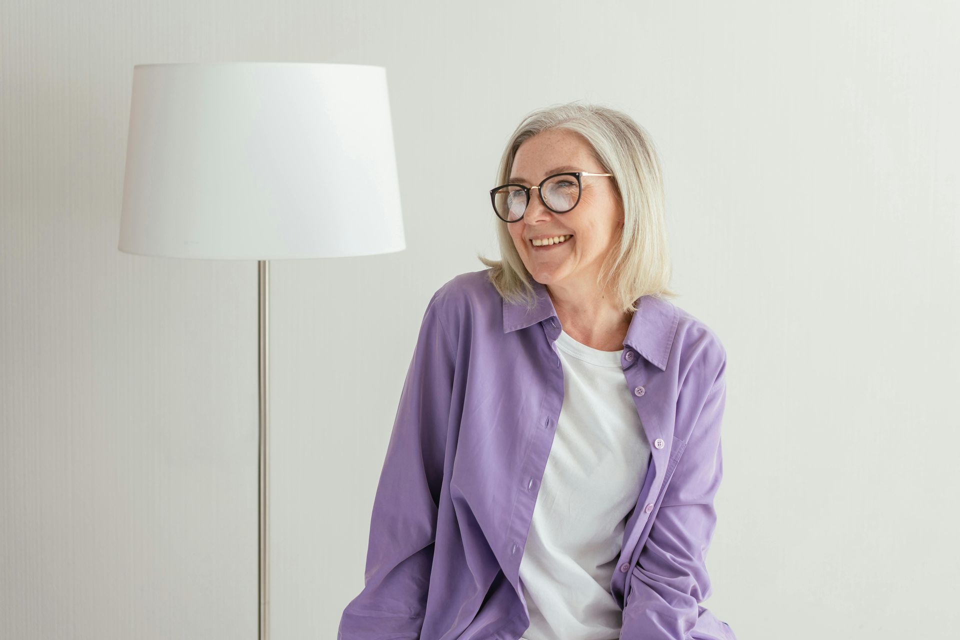 An older woman wearing glasses and a purple shirt is sitting next to a lamp.