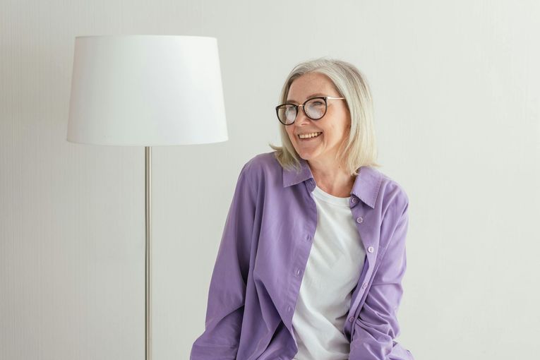 An older woman wearing glasses and a purple shirt is sitting next to a lamp.