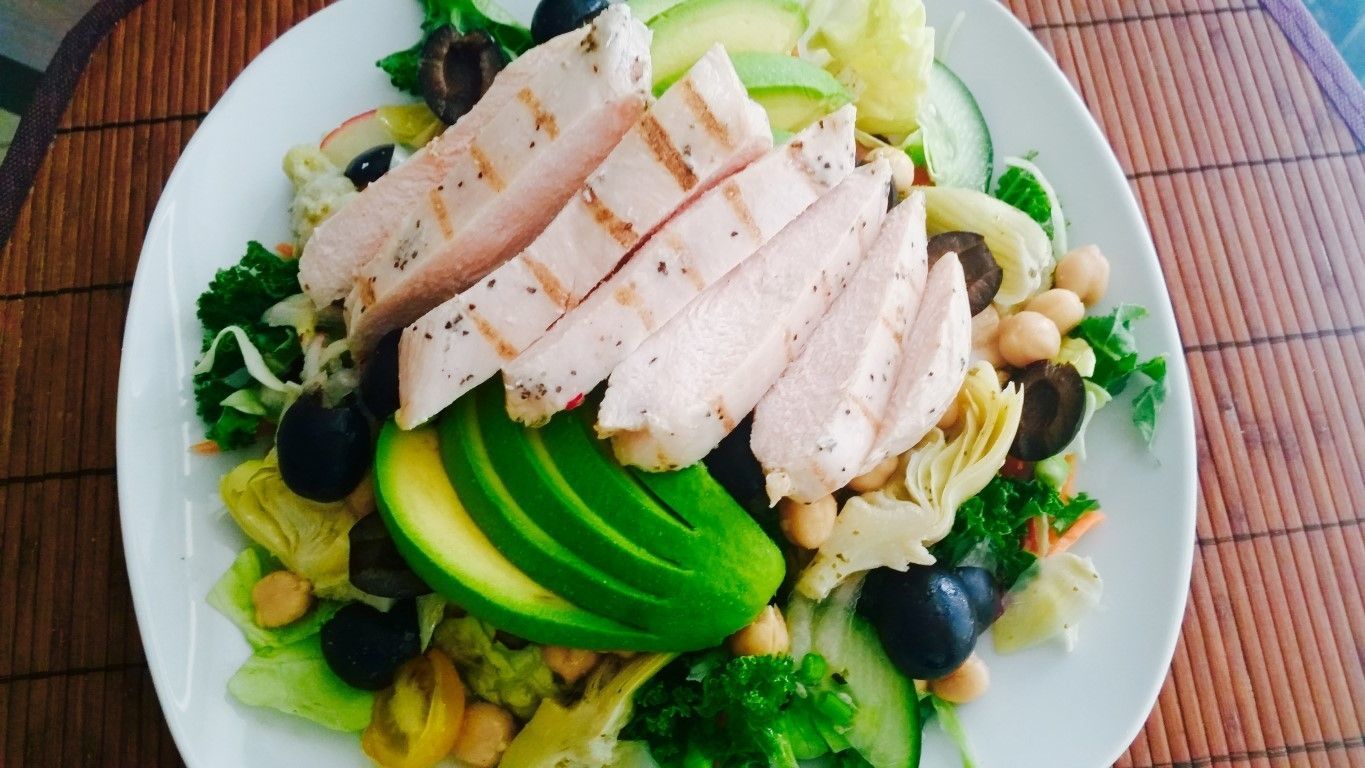 A white plate topped with a salad with chicken , avocado , olives and chickpeas.