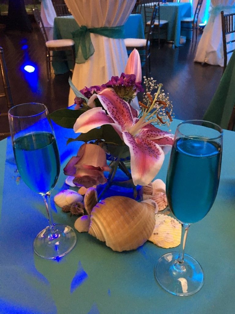 Two glasses of blue liquid are on a table with seashells and flowers.