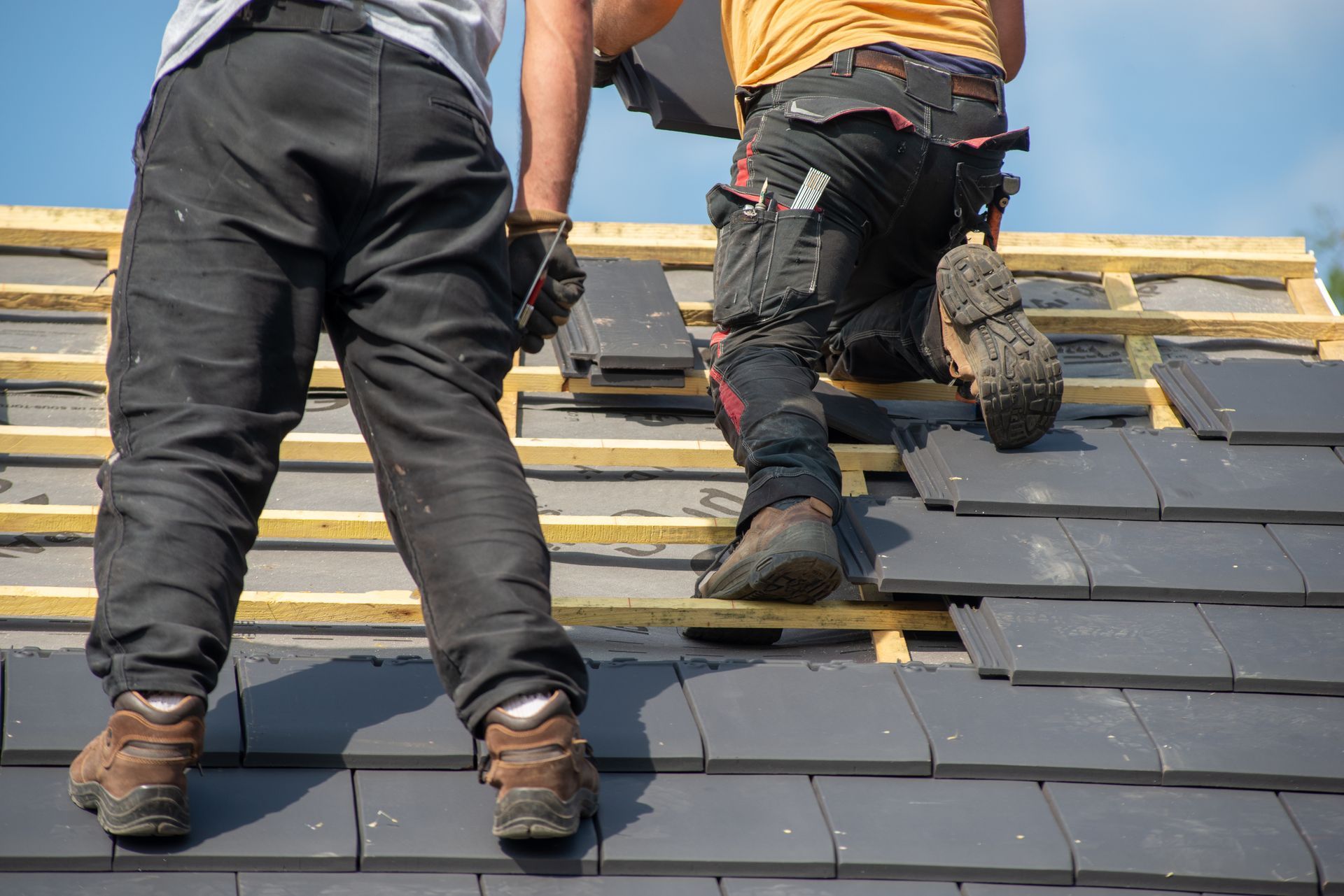 Two workers in work pants and boots installing gray tiles onto wooden roof framework.