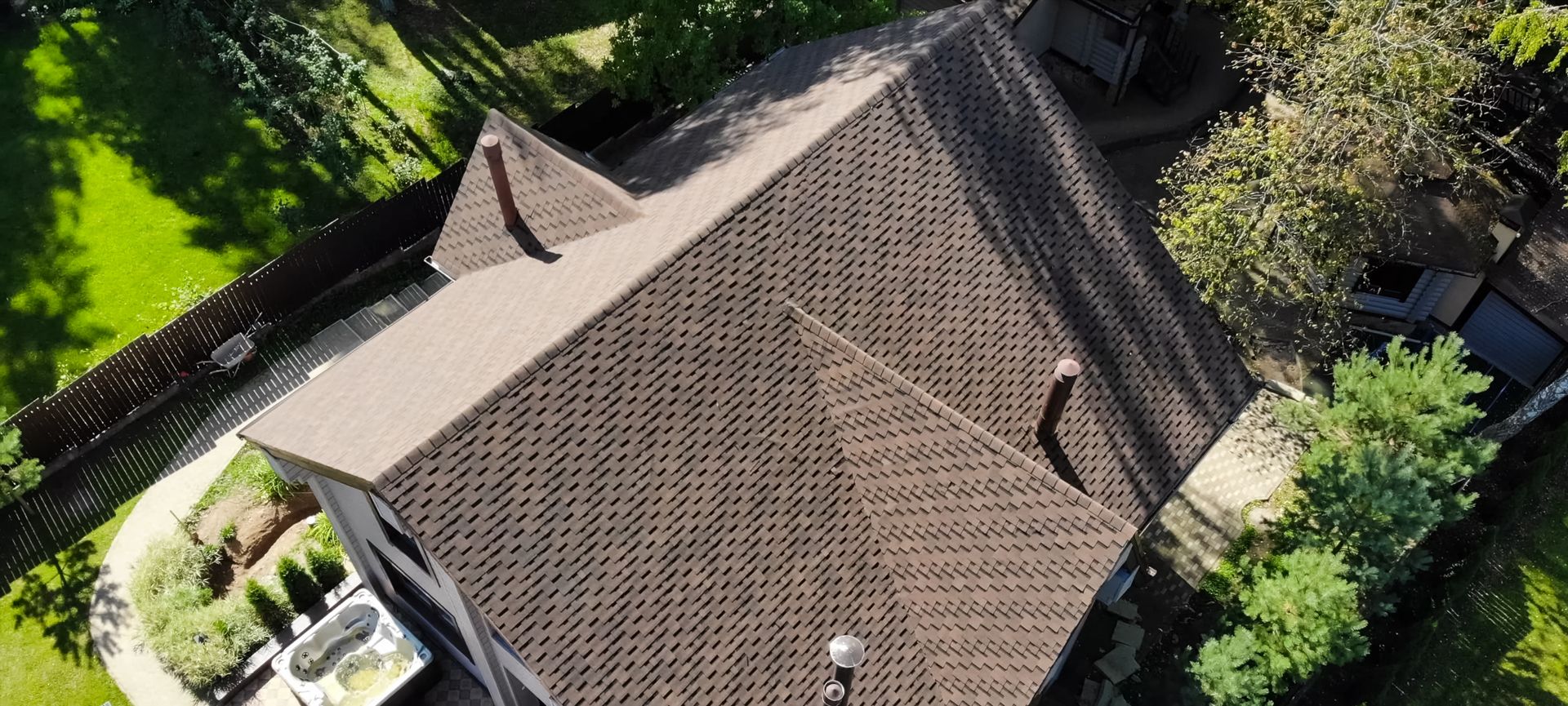 Aerial view of a brown shingled house roof surrounded by green trees and yard.