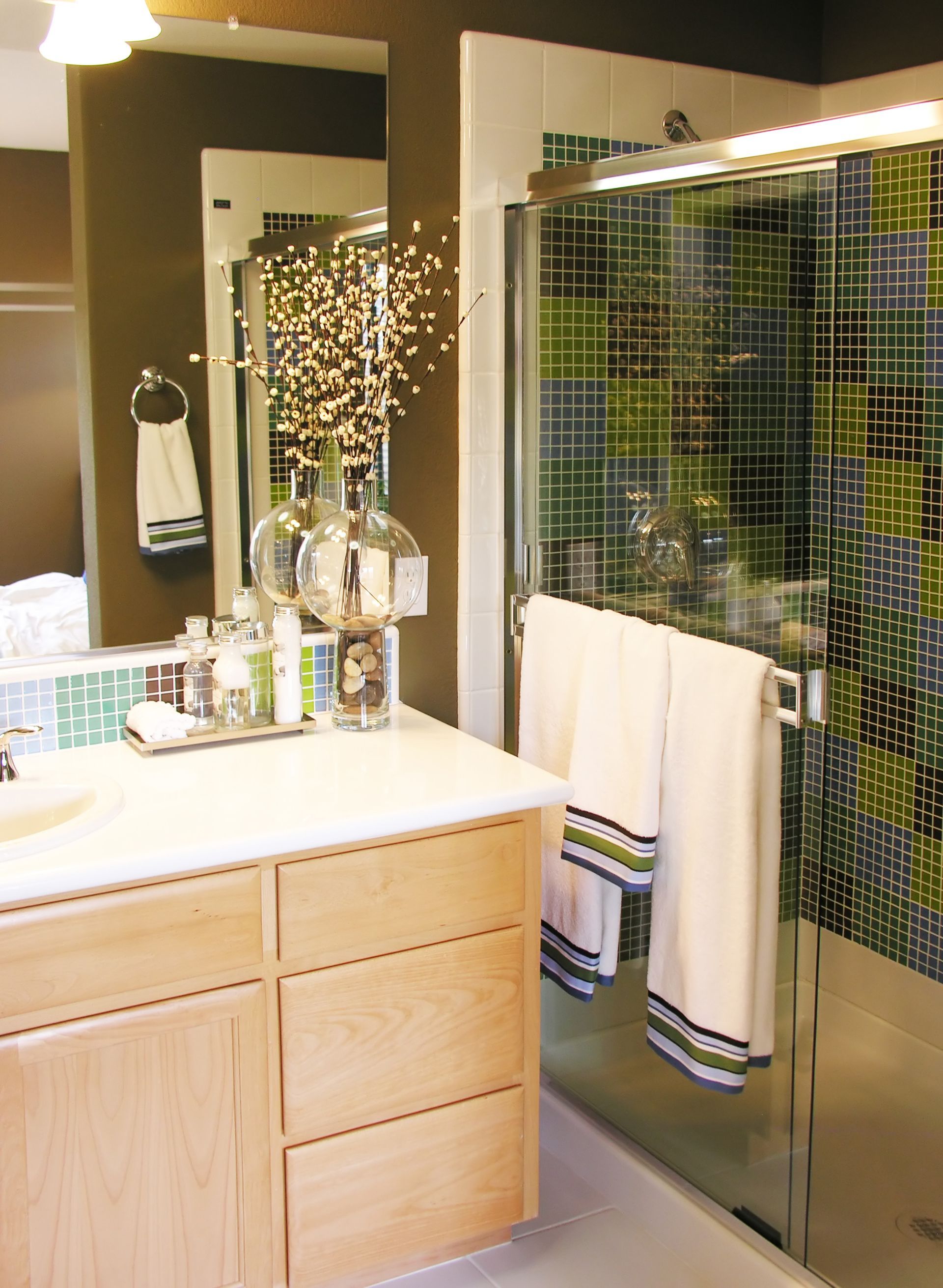 Modern bathroom vanity with mirror, bottles, towel rack, and glass shower enclosure with mosaic tile accents