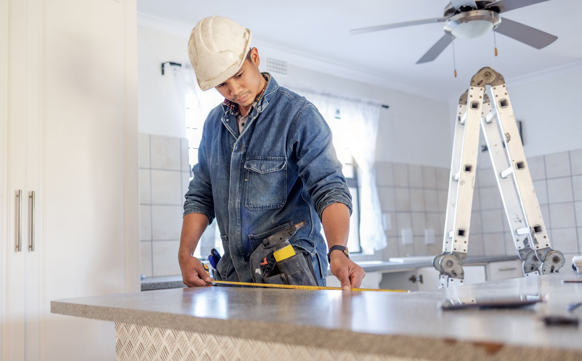 A construction worker in a white hard hat measures a kitchen countertop with a tape measure near a folding ladder.