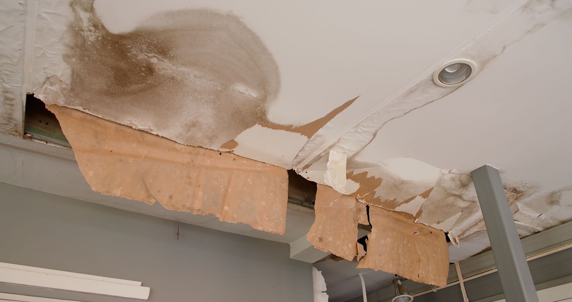 Water-damaged ceiling with peeling paint and collapsed drywall near a light fixture and shelving unit