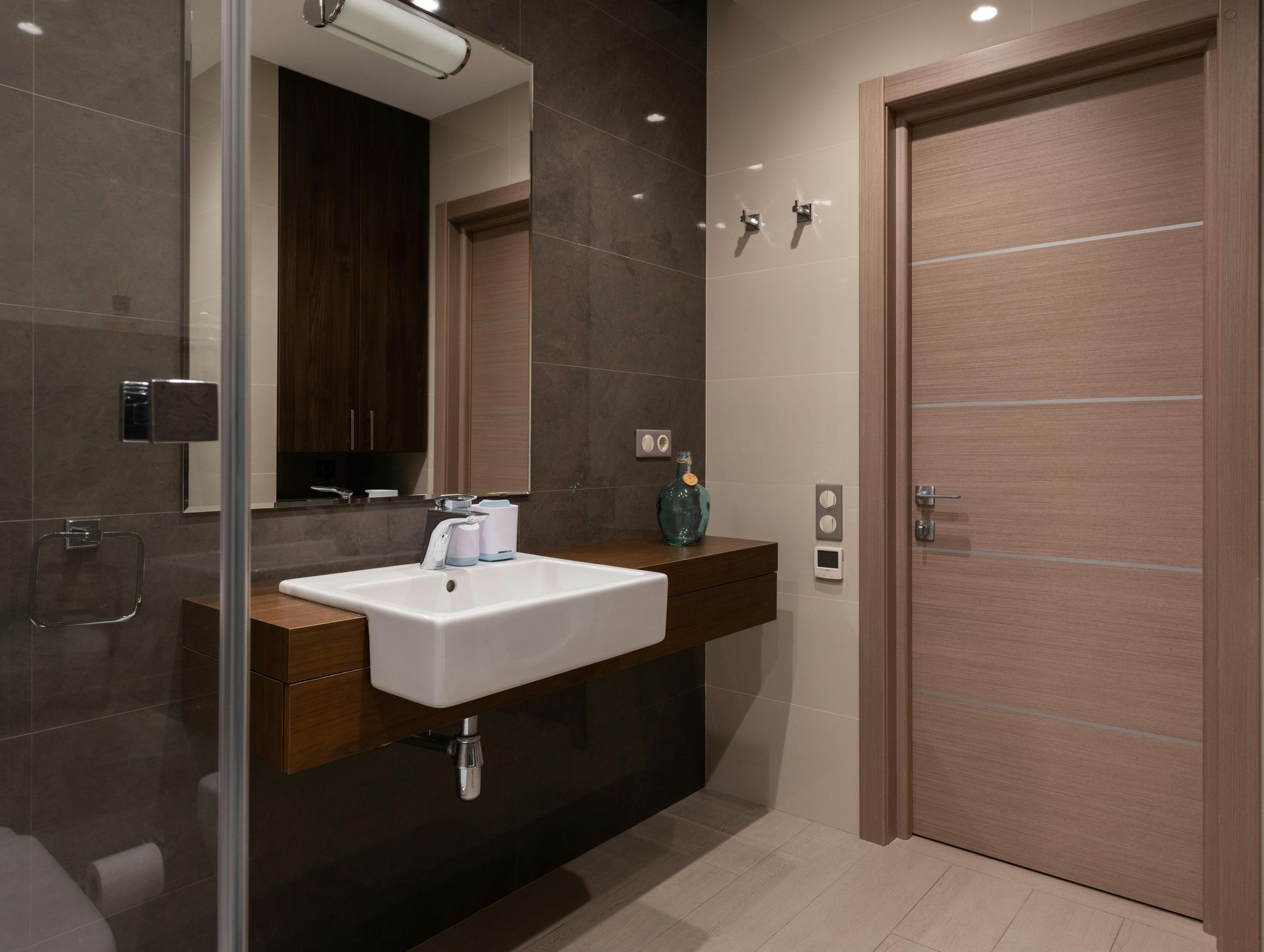Modern bathroom with glass shower, wall-mounted sink, mirror, and wooden door