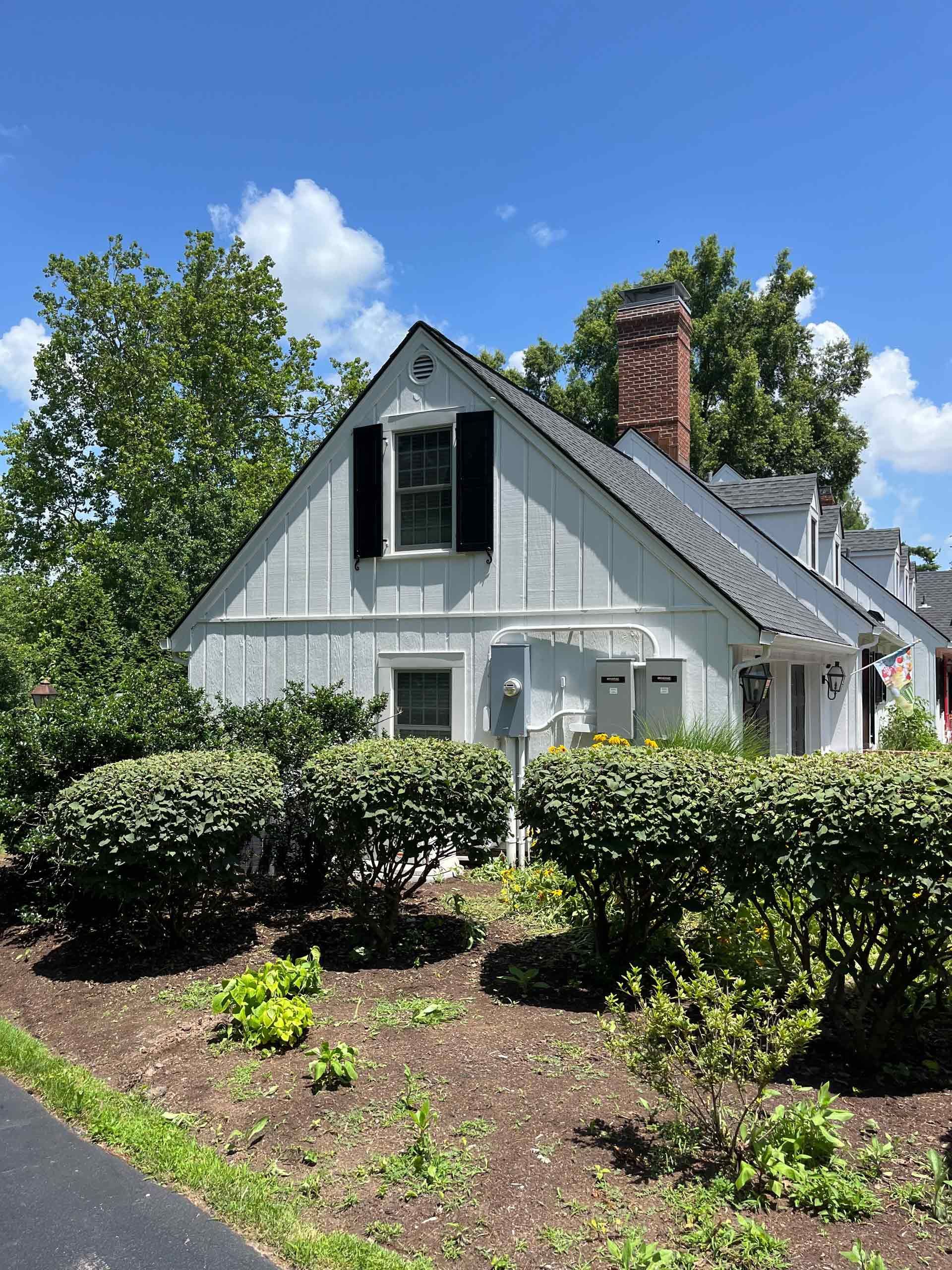 A white house with black shutters is surrounded by bushes and trees.