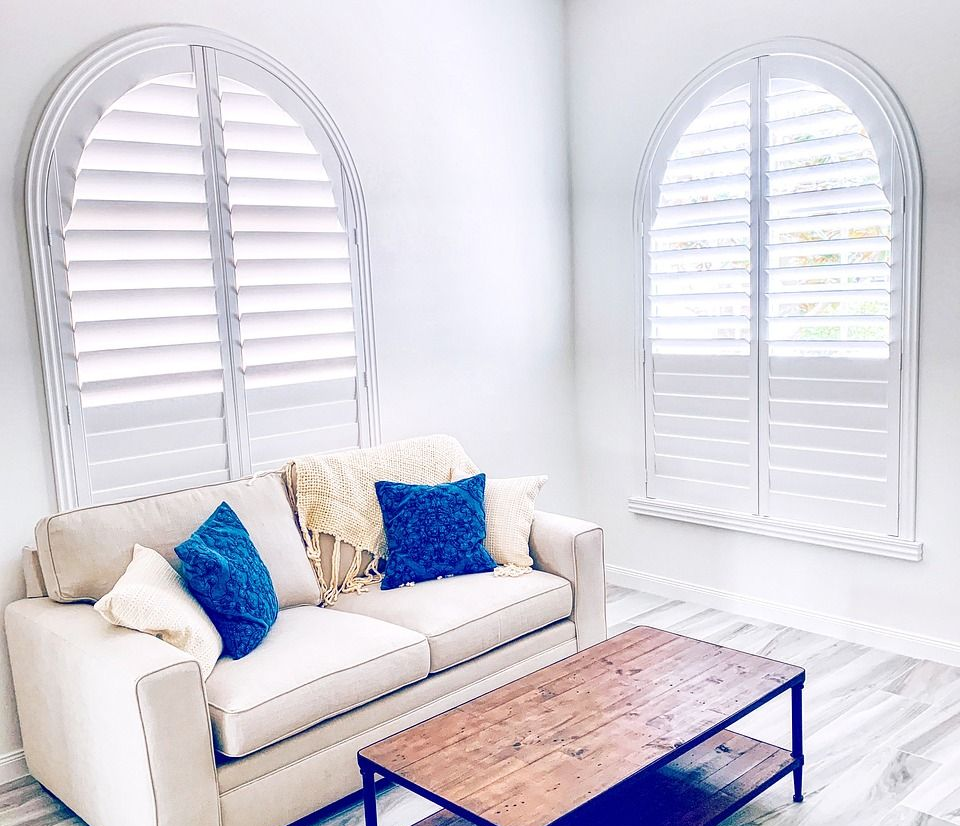 Living room with arched windows, white shutters, beige couch, blue pillows, and wooden coffee table.