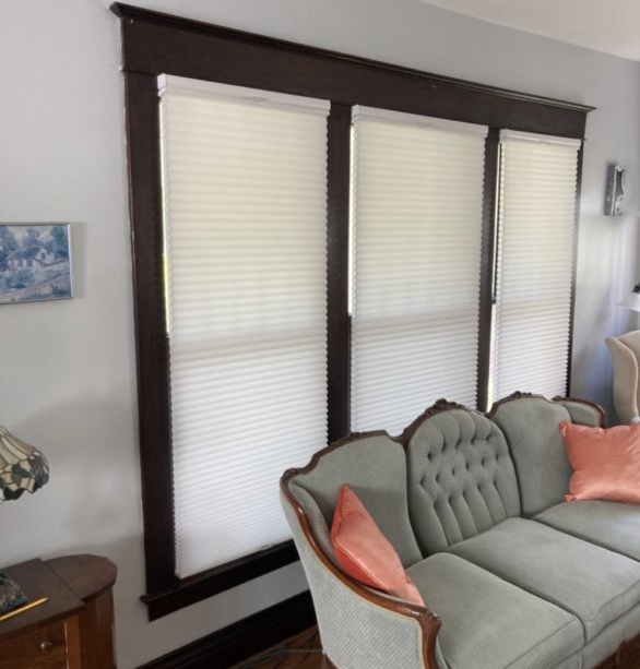 Three-pane window with white blinds, framed in dark wood. A gray couch with peach pillows sits below.
