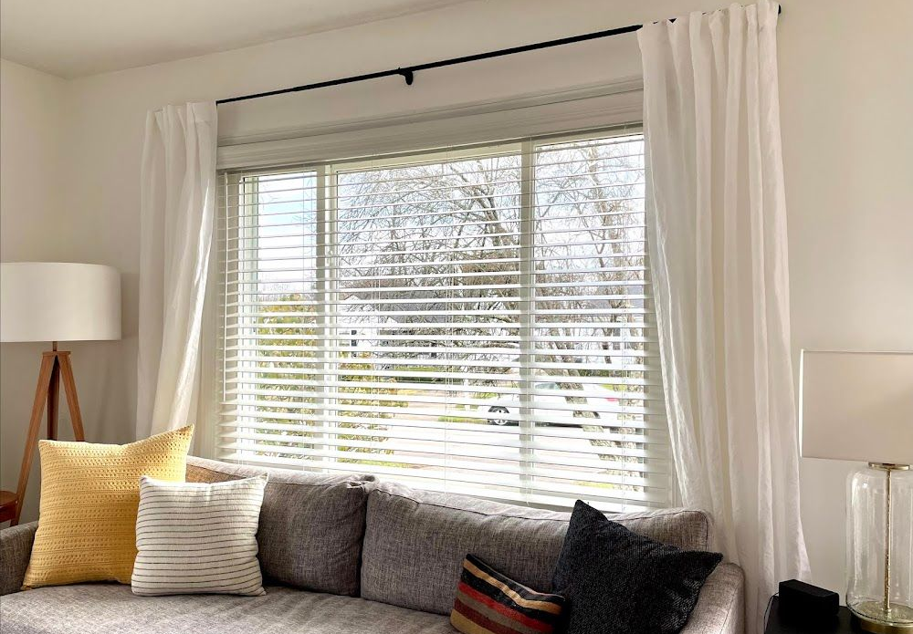 Living room with a window, white curtains, gray couch, pillows, and lamps.