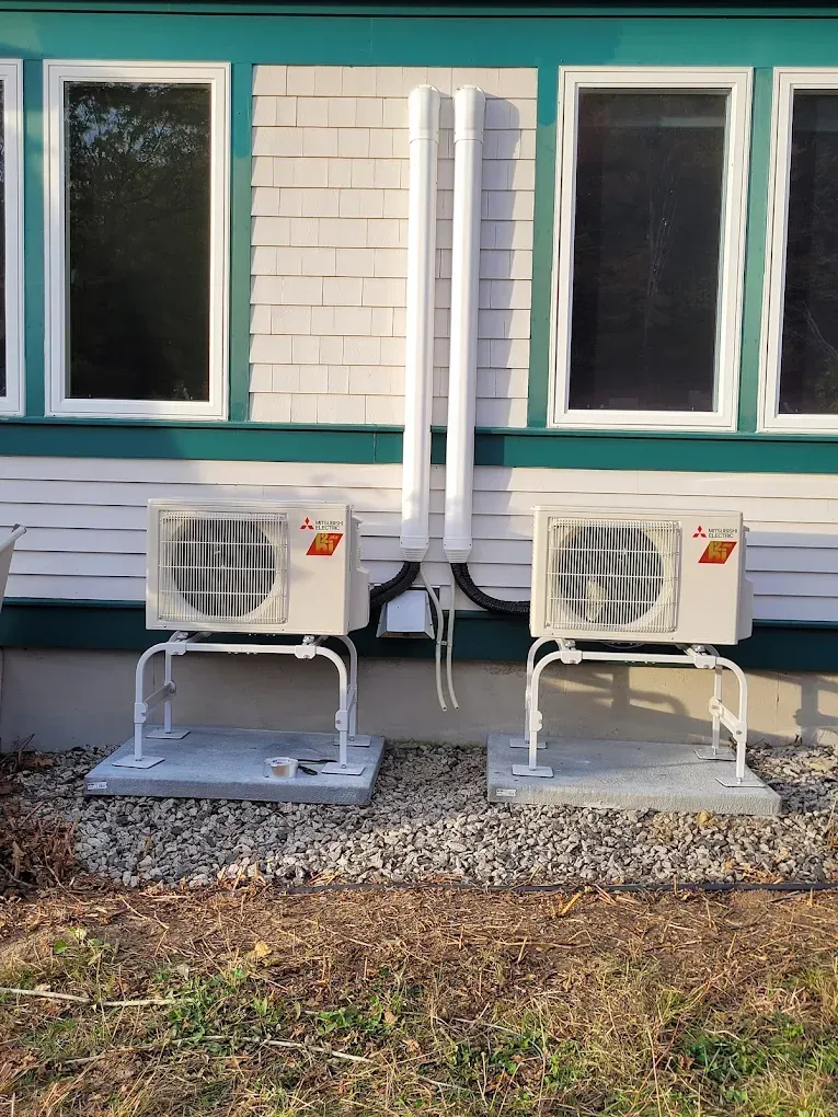 Two white Mitsubishi mini-split outdoor units sitting on concrete pads against a white wall with two vertical pipes.