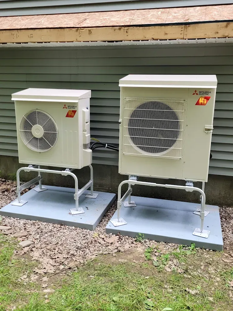 Two beige Mitsubishi outdoor HVAC units on metal stands over concrete pads against a gray siding wall.