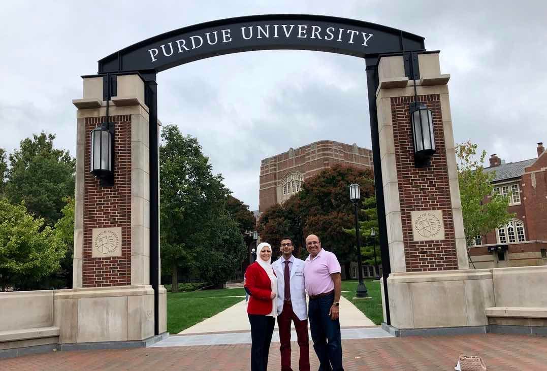 Three people standing under the Purdue University gate arch outdoors.