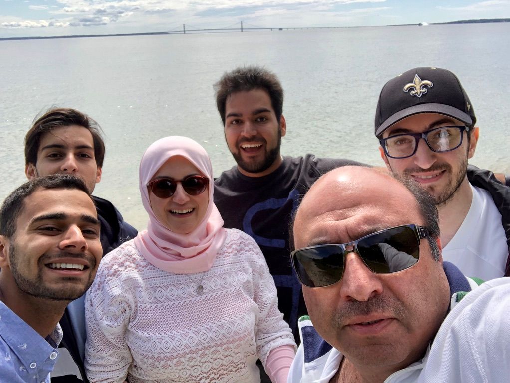 Group of six smiling people posing for a selfie by the water on a bright day.