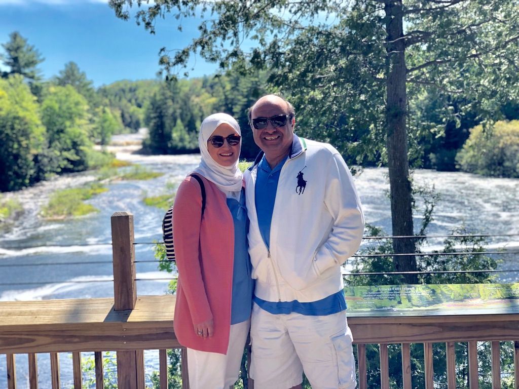 Couple standing on a wooden deck by a rushing river, with trees and a bright blue sky behind them.