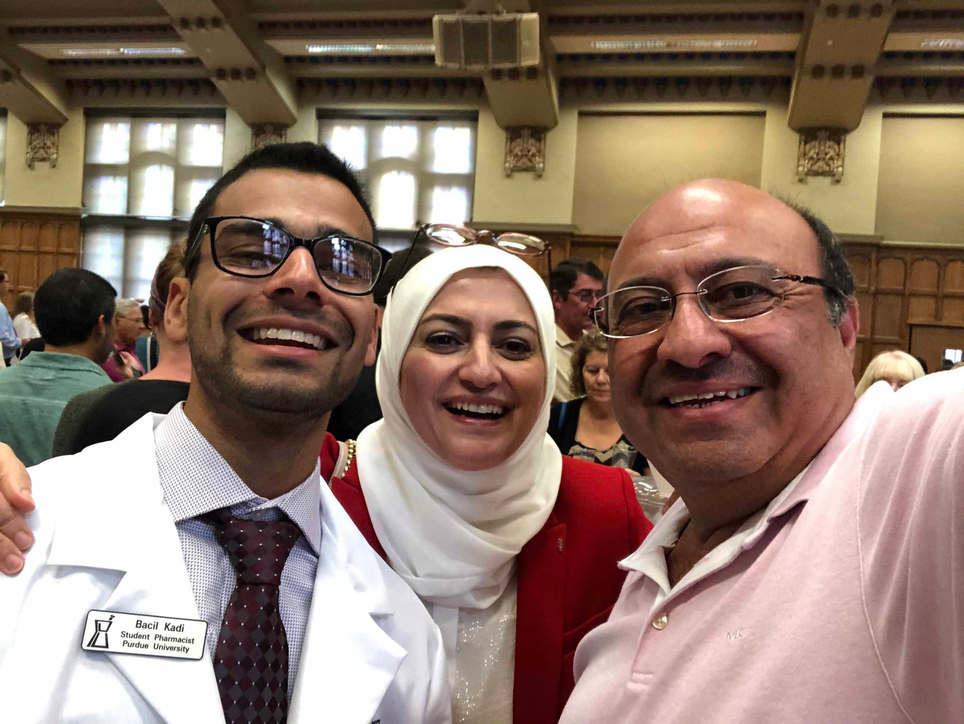 Three smiling people posing together indoors at an event, one in a white coat, one in a headscarf, one in a pink shirt